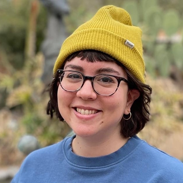 Photo of Aubrey outdoors, smiling, wearing glasses and a mustard yellow beanie.